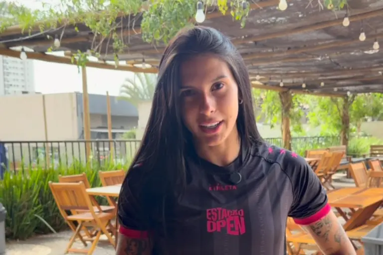 Ana Castela com cabelos longos e escuros, vestindo uma camisa esportiva preta com detalhes em rosa e a estampa "Estação Open". Ao fundo, um ambiente aberto com mesas de madeira e luzes decorativas penduradas.