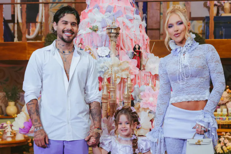 Zé Felipe, vestido com camisa branca e calça lilás, segura a mão da filha Maria Flor, que usa um vestido lilás e detalhes florais no cabelo. À direita, Virginia Fonseca veste um conjunto de renda lilás com gola alta e mangas longas. Os três sorriem para a câmera, posicionados em frente a um grande bolo de aniversário temático decorado em tons de rosa, babados e torres de castelo.