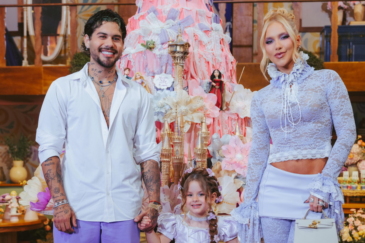 Zé Felipe, vestido com camisa branca e calça lilás, segura a mão da filha Maria Flor, que usa um vestido lilás e detalhes florais no cabelo. À direita, Virginia Fonseca veste um conjunto de renda lilás com gola alta e mangas longas. Os três sorriem para a câmera, posicionados em frente a um grande bolo de aniversário temático decorado em tons de rosa, babados e torres de castelo.