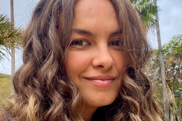 Lua Blanco sorri ao ar livre com cabelo cacheado loiro e castanho, vestindo blusa azul e tendo palmeiras e área verde ao fundo