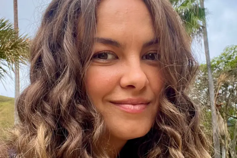 Lua Blanco sorri ao ar livre com cabelo cacheado loiro e castanho, vestindo blusa azul e tendo palmeiras e área verde ao fundo