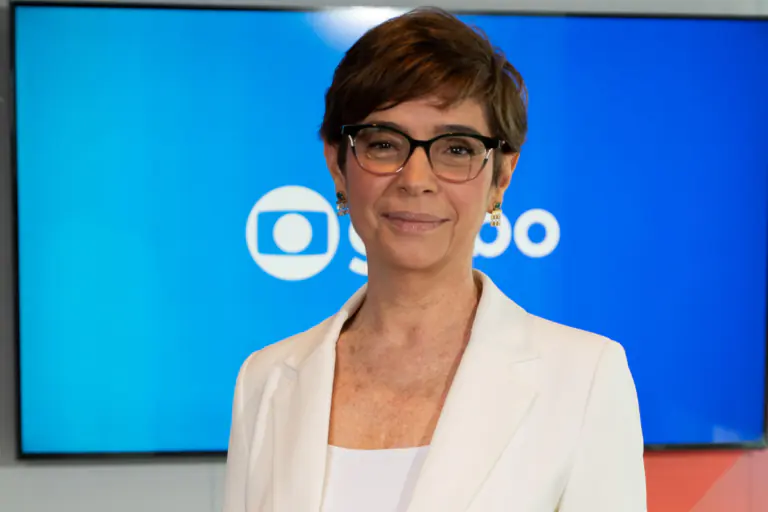 Renata Lo Prete, com cabelos curtos castanhos e óculos de grau, sorri levemente olhando de frente. Ela veste um blazer e calça brancos sobre uma blusa da mesma cor. Ao fundo, há um telão azul com o logotipo da TV Globo e painéis geométricos em laranja e vermelho.
