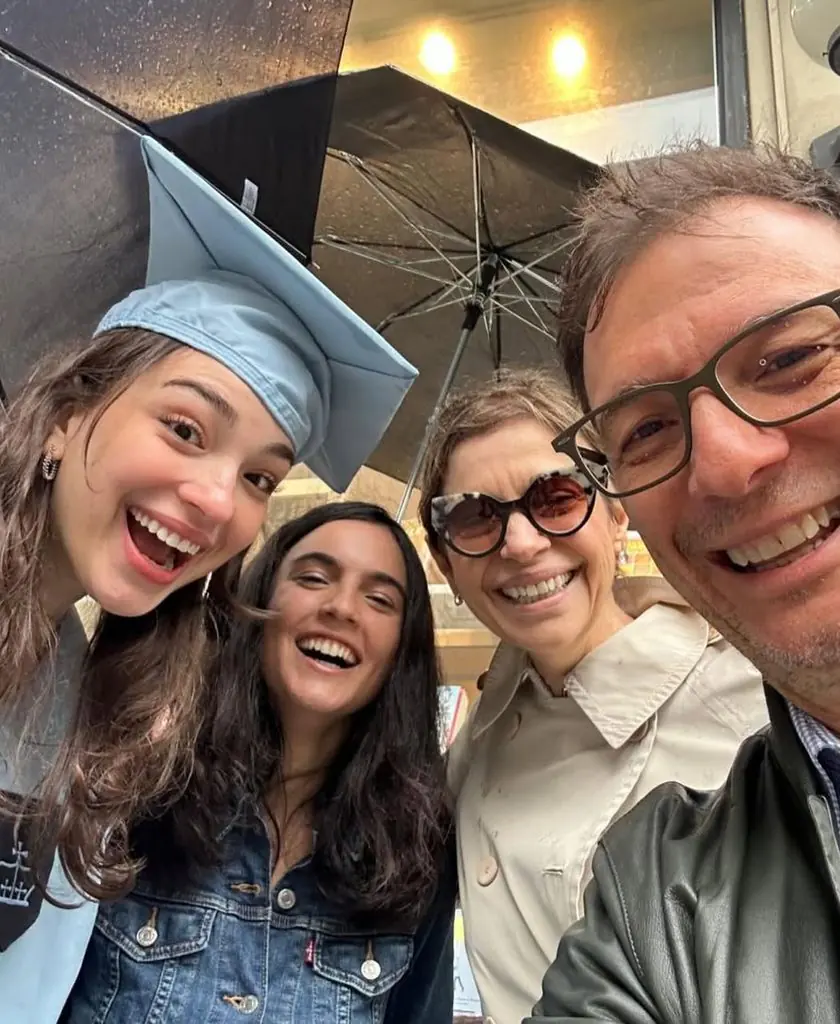 Quatro pessoas sorrindo para uma selfie ao ar livre sob guarda-chuvas pretos. À esquerda, uma jovem usa capelo de formatura azul-claro. Ao centro, outra jovem com jaqueta jeans sorri abertamente. À direita, a jornalista Renata Lo Prete, de óculos escuros e casaco claro, está ao lado do marido, Melchiades Filho, que veste jaqueta escura e óculos de grau.