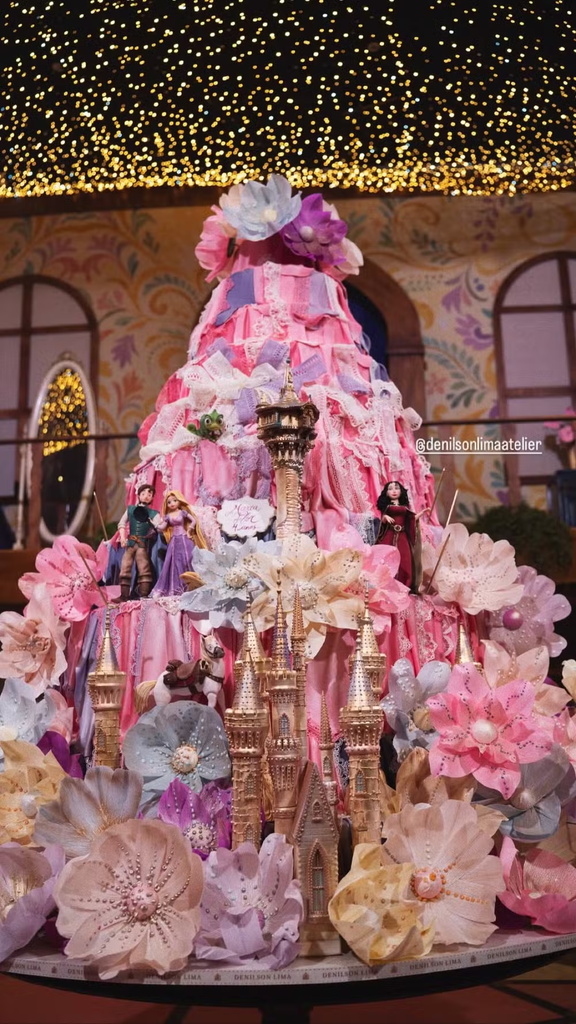 Bolo de aniversário temático alto, decorado com babados em tons de rosa e roxo, grandes flores, torres de castelo douradas na base e miniaturas de personagens de contos de fadas, iluminado por um teto de luzinhas ao fundo.