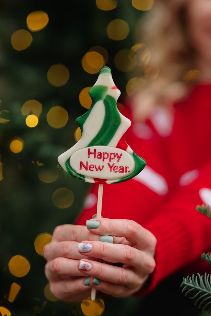 Hand holding a tree-shaped lollipop with Happy New Year text. Festive lights background.